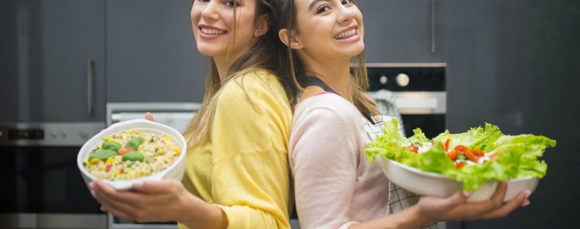 Two smiling women hold vibrant salads in a kitchen setting, showcasing fresh culinary creations.
